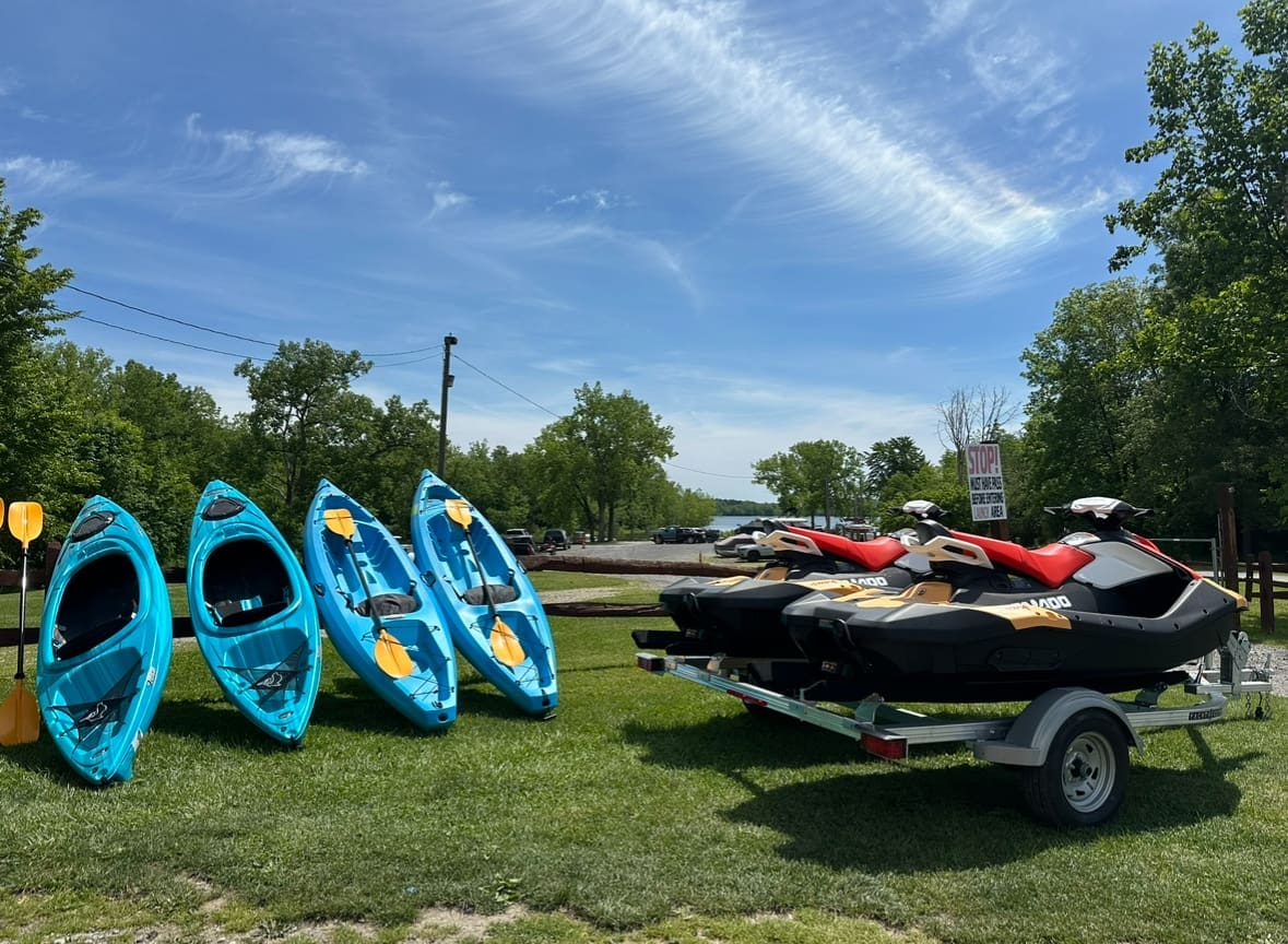 Four blue kayaks lined up along a stand. Nearby, two black and red sea doo jet skis are on the right.